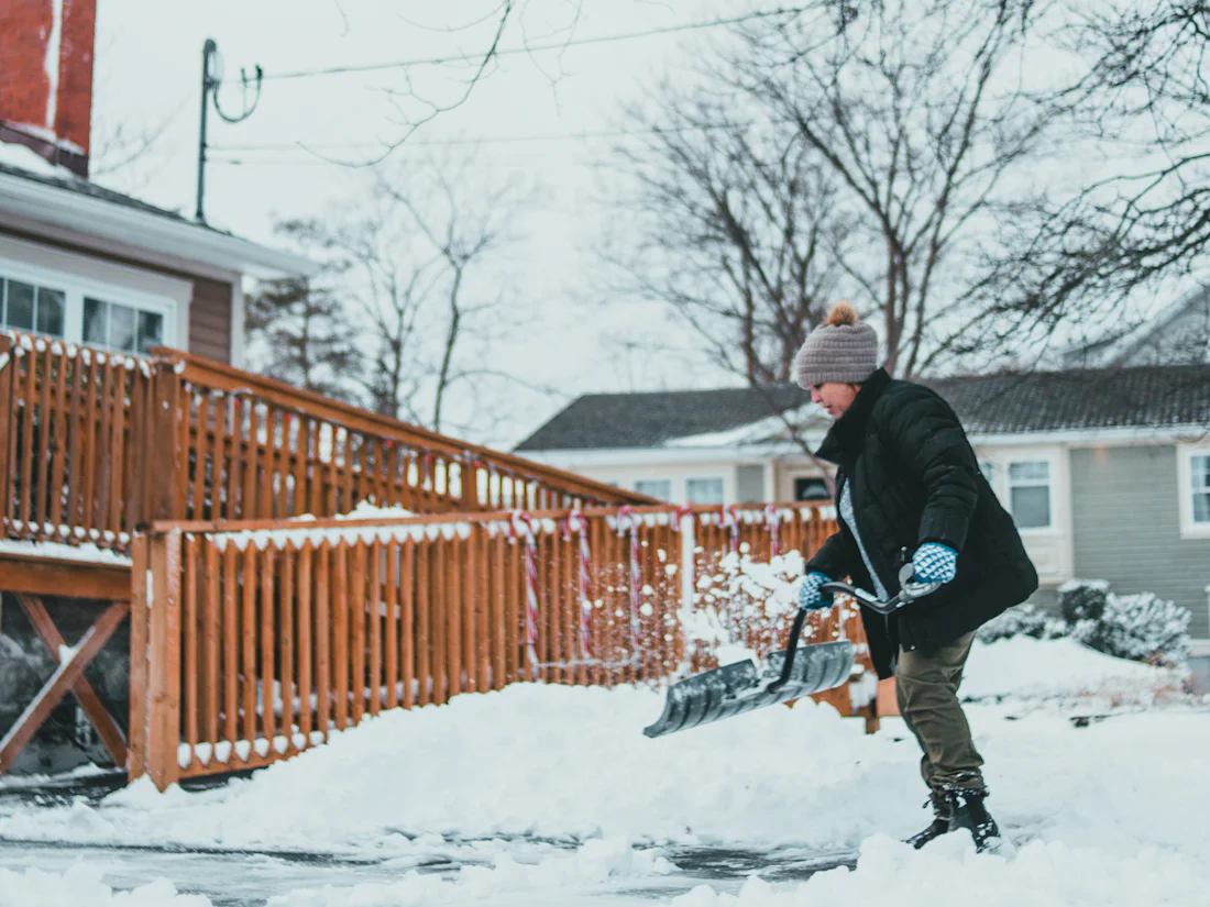 Mann räumt mit einer Schneeschaufel eine verschneite Einfahrt vor einem Holzzaun